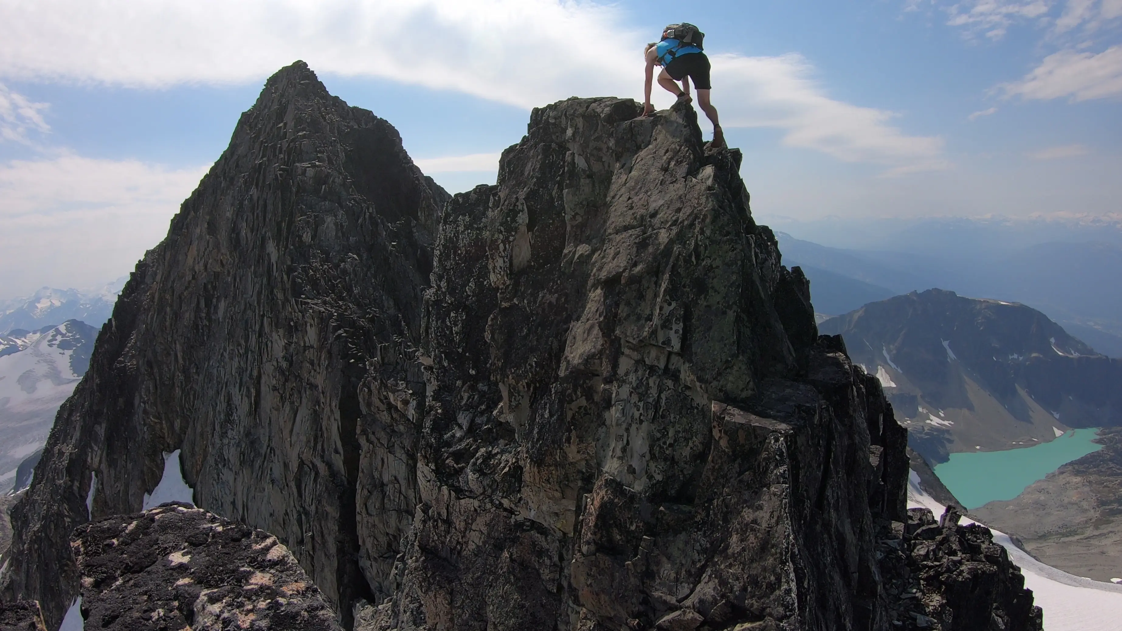 A climber ascending one of the final technical steps to reach the summit of Mount Weart and complete the Armchair Traverse with lakes and glaciers far below.