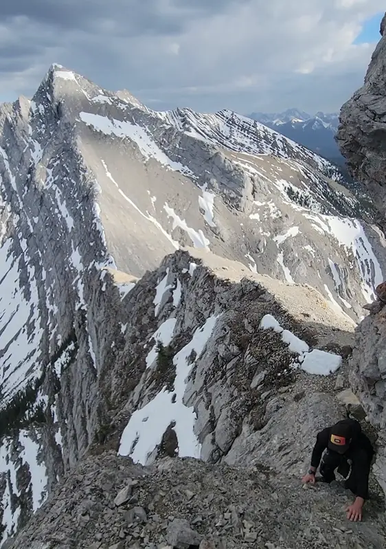 A climber on an exposed ledge on Mount McGillivray to reach the summit with Skogan North Peak in the background.