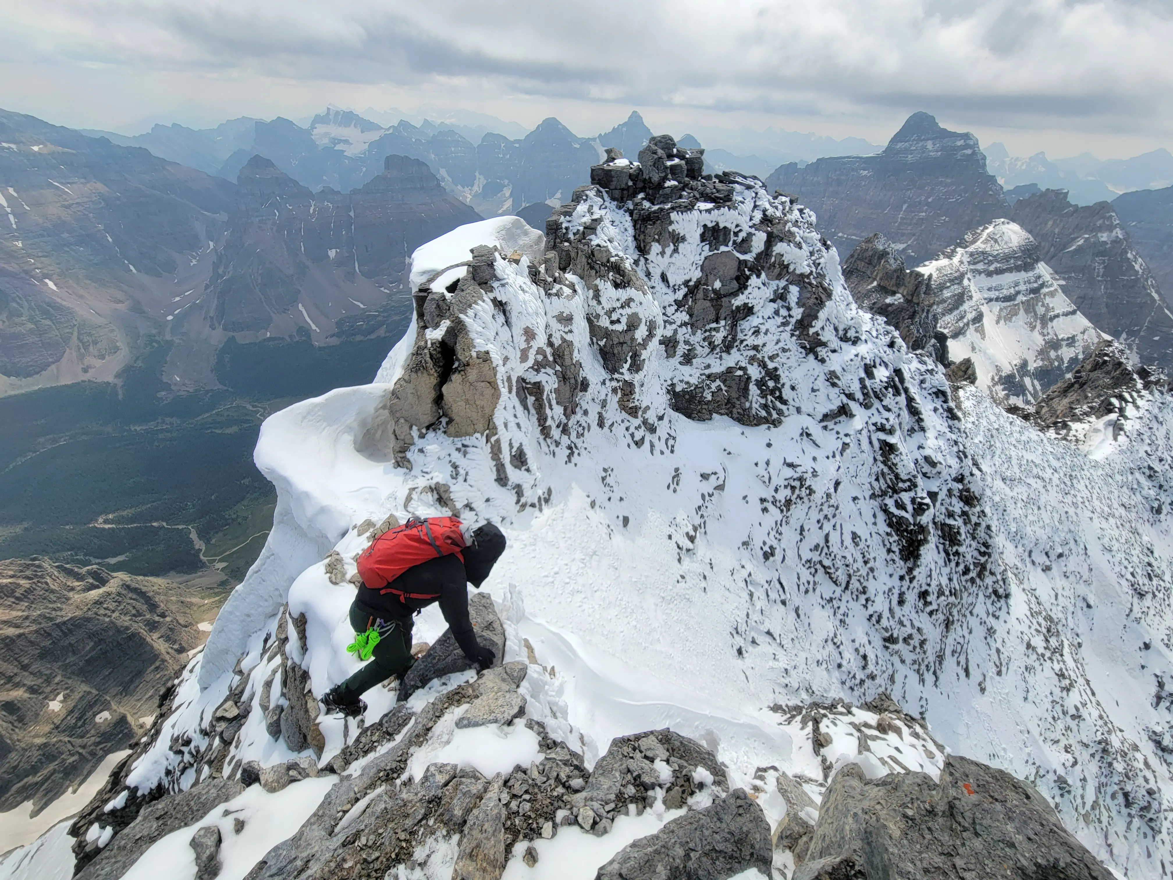 A climbed descending the exposed and snowy ridge of Mount Lefroy with peaks like Mount Fay, Mount Deltaform, and Hungabee Mountain along the horizon.