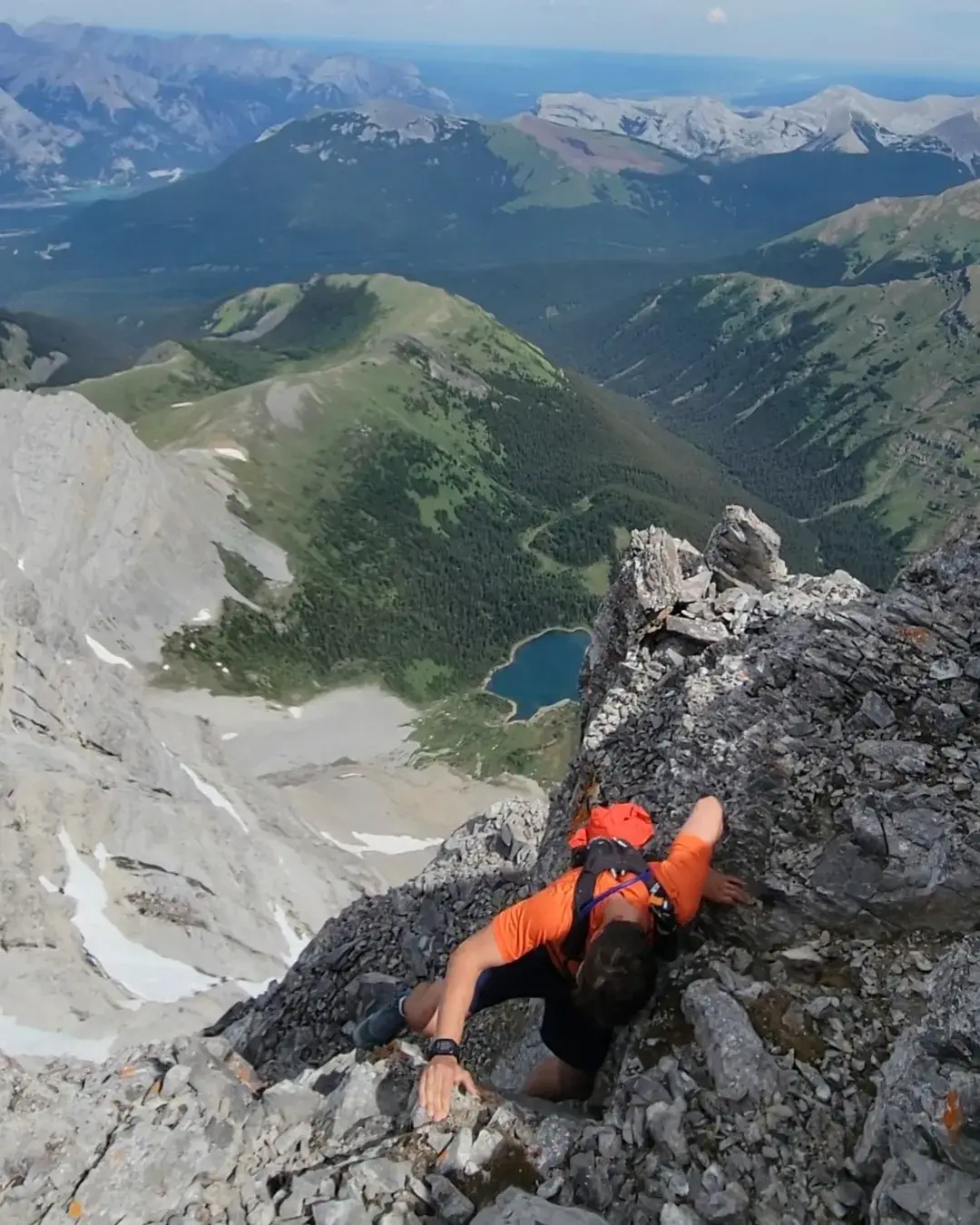 A climber ascending the crux of the Lougheed Traverse on the second peak with a sizable drop below and a small alpine lake.