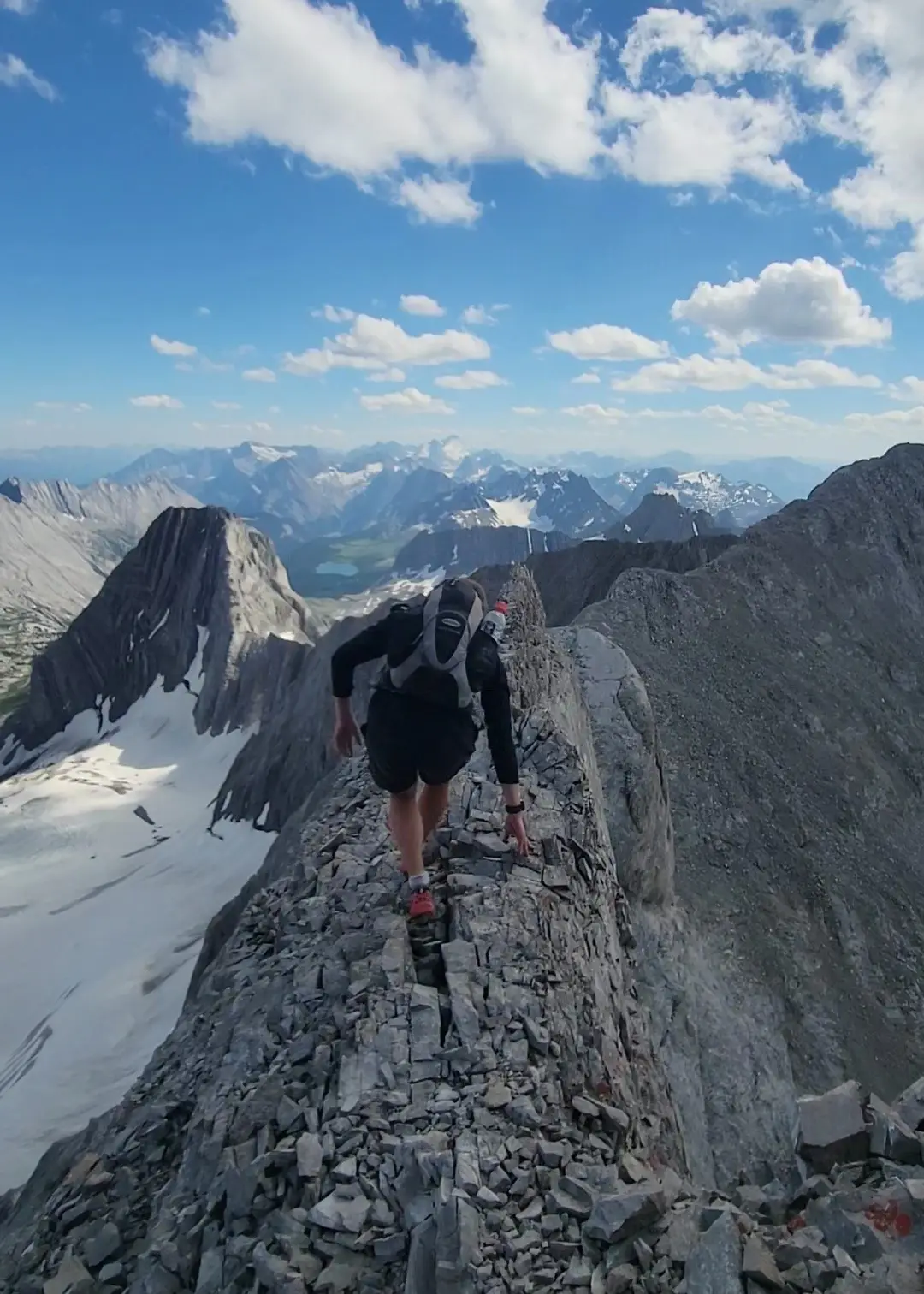 A climber crosses the exceedingly exposed ridge of Mount French nicknamed The Guillotine with impressive glacier and lakes below and peaks on the horizon.