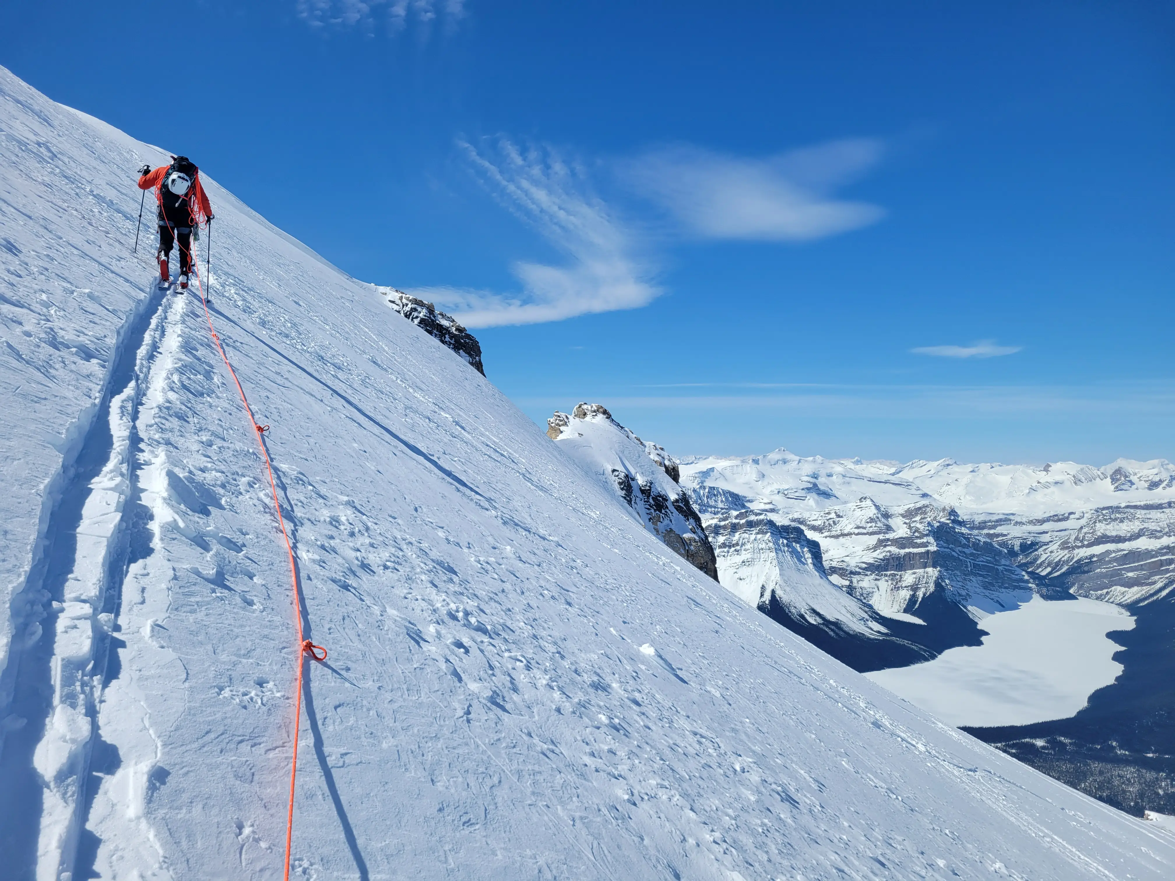 Ascending Mount Hector on skis, roped up for glacier travel with Hector Lake visible far below to the right under clear blue skies