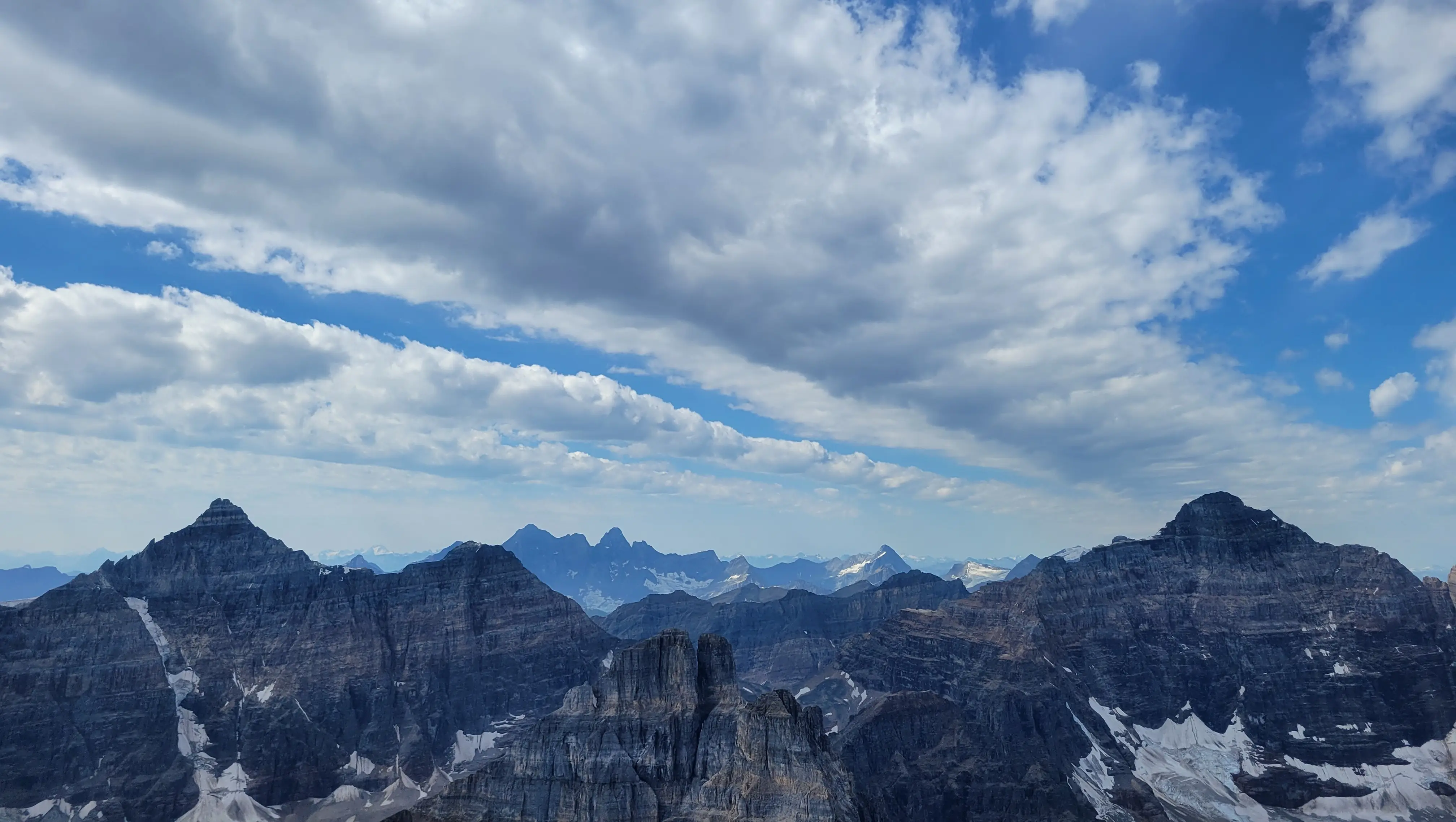 Panoramic view from Mount Temple showing rugged peaks of the Canadian Rockies under a sky with scattered clouds