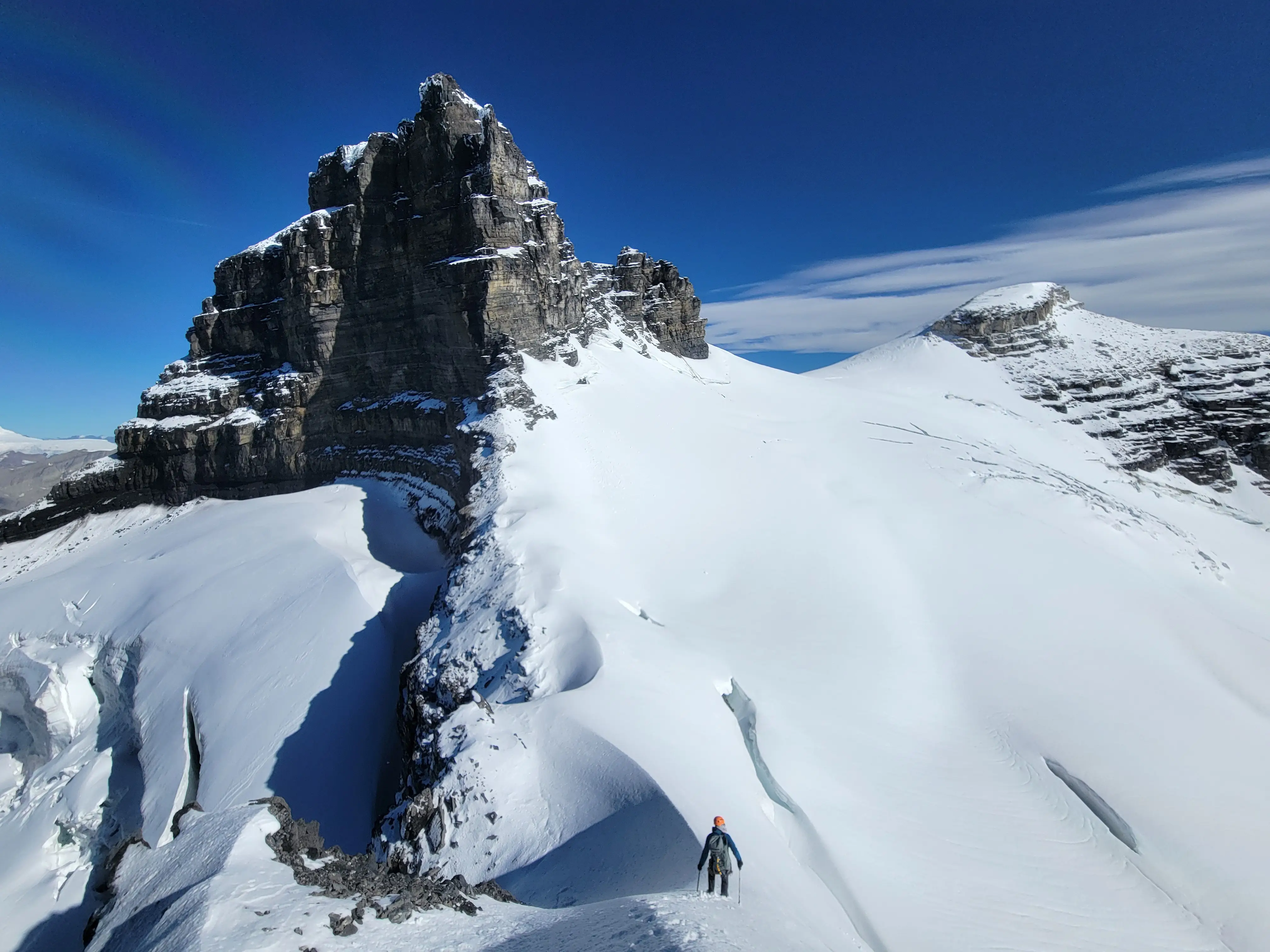 Climber descending the exposed north ridge of Lyell 4 with crevasses below and Lyell 3 visible ahead under a blue sky in the remote Canadian Rockies.
